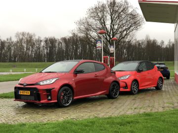 Rode Toyota GR Yaris geparkeerd bij een benzinestation met een andere auto en een grasveld op de achtergrond op een bewolkte dag.