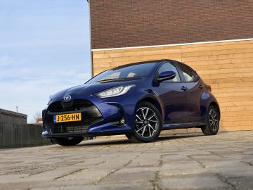 Een blauwe Toyota Yaris-benzine geparkeerd op de stoep voor een houten hek onder een heldere hemel.
