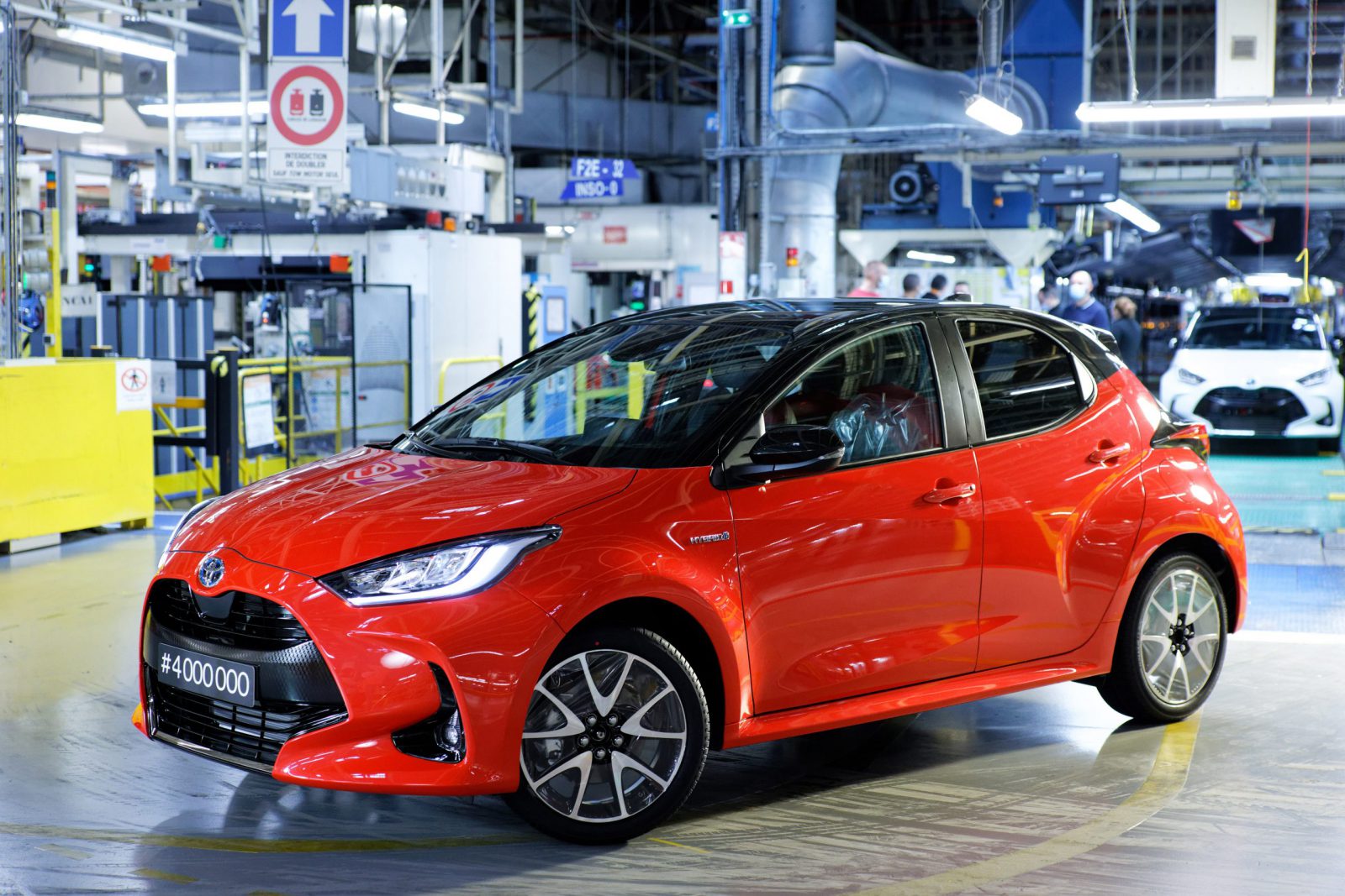 Een rode Toyota Yaris sedan geparkeerd in een helder verlichte autofabriek met verschillende machines en activiteiten op de achtergrond.