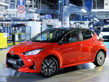 Een rode Toyota Yaris sedan geparkeerd in een helder verlichte autofabriek met verschillende machines en activiteiten op de achtergrond.