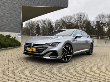 Een zilveren Volkswagen Arteon Shooting Brake geparkeerd op een betonnen stoep nabij een modern gebouw, onder een grote overhangende structuur, op een bewolkte dag.