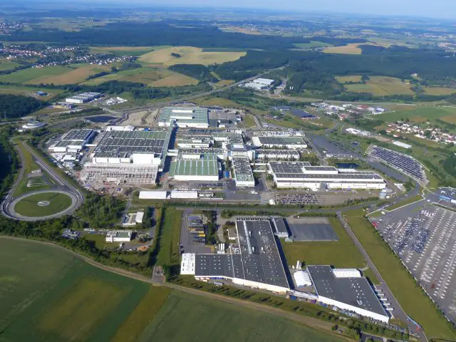 Luchtfoto van een groot industrieel complex van INEOS Automotive met meerdere gebouwen, parkeerplaatsen en omgeven door groene velden.