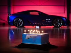 Een verjaardagstaart met brandende kaarsen in de vorm van het Lexus LFA-logo, voor een blauwe Lexus-sportwagen onder rode verlichting.