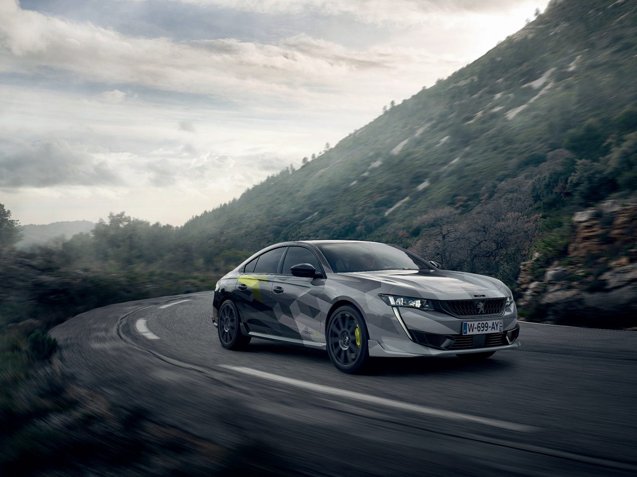 Een gecamoufleerde Peugeot 508 PSE die onder een bewolkte hemel te hard rijdt op een bochtige bergweg.