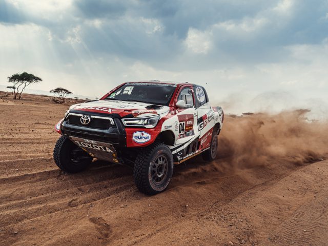 Een Dakar-rallyauto met meerdere sponsorlogo's doet stof opwaaien tijdens het racen op een onverharde baan in een woestijnlandschap onder een gedeeltelijk bewolkte hemel.
