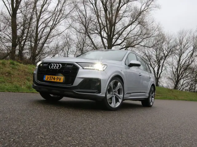 Een zilveren Audi Q7 SUV met koplampen staat geparkeerd op een verharde weg naast grasachtig terrein en bladerloze bomen op een bewolkte dag.