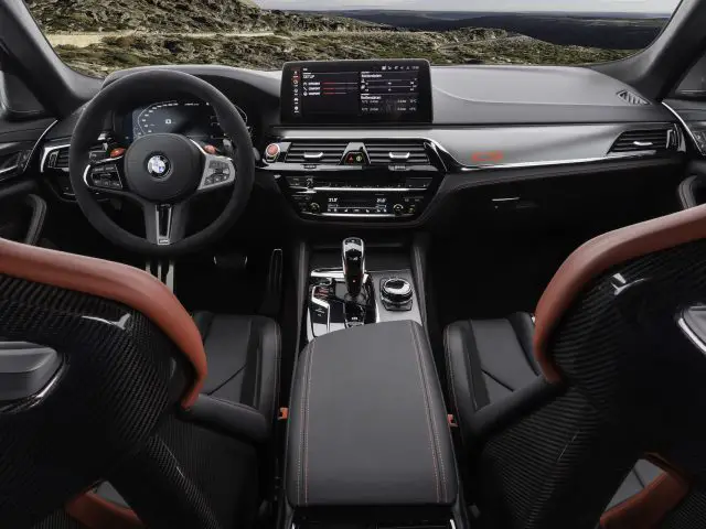 Binnenaanzicht van een hoogwaardige BMW M5 CS met een modern dashboard, touchscreen-display en strakke zwart-rode lederen stoelen. De versnellingspook, het stuur en diverse bedieningselementen zijn ook zichtbaar.