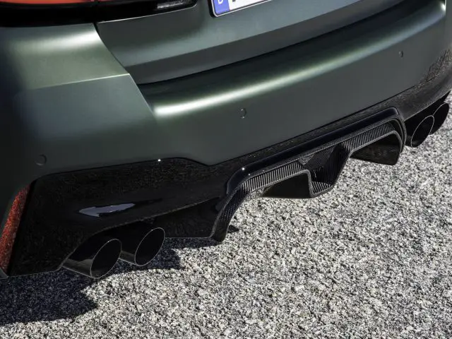 Close-up van de achterbumper van een BMW M5 CS, met aan elke kant een zwarte diffuser en dubbele uitlaatpijpen. De achtergrond bestaat uit grind.