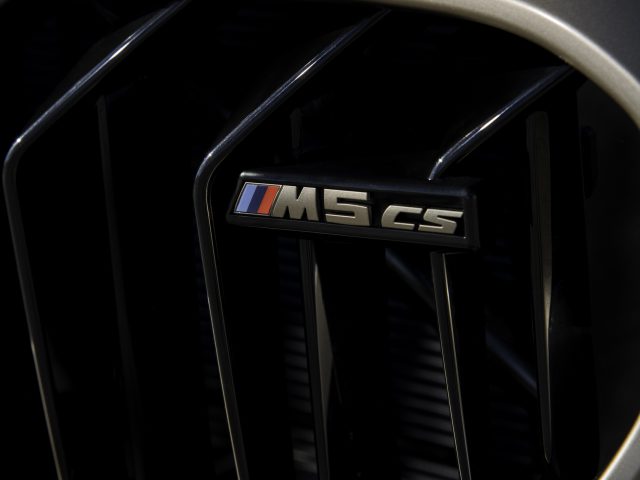 Close-up van een BMW M5 CS-grille met een embleem met de tekst "M5 CS" en een klein driekleurenlogo. De grille bestaat uit verticale zwarte lamellen, wat doet denken aan een high-performance voertuig.