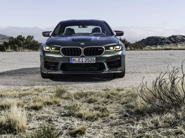Een vooraanzicht van een zwarte BMW M5 CS-auto geparkeerd op een verlaten, rotsachtig landschap met struiken op de voorgrond en heuvels op de achtergrond.
