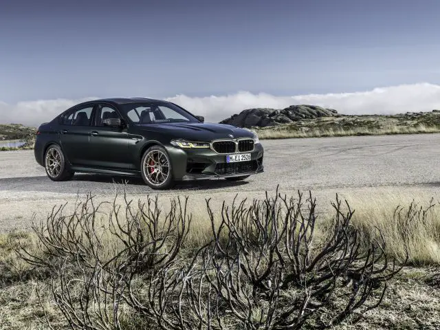 Een donkergroene BMW M5 CS sedan staat geparkeerd op een verlaten weg met droge vegetatie op de voorgrond en rotsachtig terrein op de achtergrond onder een heldere hemel.