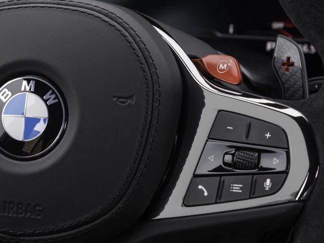 Close-up van een BMW-stuur met het logo, de bedieningsknoppen en een rode "M2"-knop, die doet denken aan de krachtige BMW M5 CS.