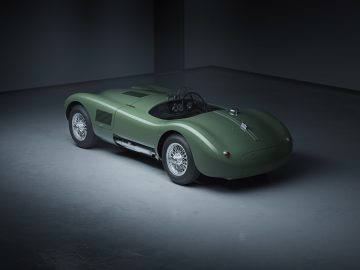 Een vintage groene Jaguar C-type raceauto met een strak, aerodynamisch ontwerp staat geparkeerd in een slecht verlichte, lege garage. De auto heeft zichtbare wielen en een laag profiel.