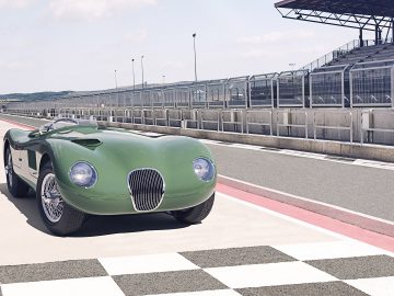 Een vintage groene Jaguar C-type cabriolet staat geparkeerd op de pitlane van een racecircuit vlakbij de startlijn, met lege tribunes en een heldere hemel op de achtergrond.
