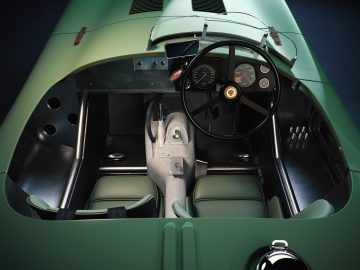 Een bovenaanzicht van het interieur van een klassieke groene Jaguar C-type raceauto met een stuur, meters en twee stoelen.