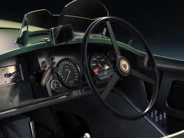 Close-up van een vintage Jaguar C-type dashboard met een groot stuur, klassieke snelheidsmeters, een gemonteerde smartphone en een zonnebril die naast de snelheidsmeter hangt.