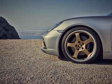 Zilveren Porsche Boxster geparkeerd op een grindweg met uitzicht op een kustlandschap, met de nadruk op het linker voorwiel en de koplamp van het voertuig.