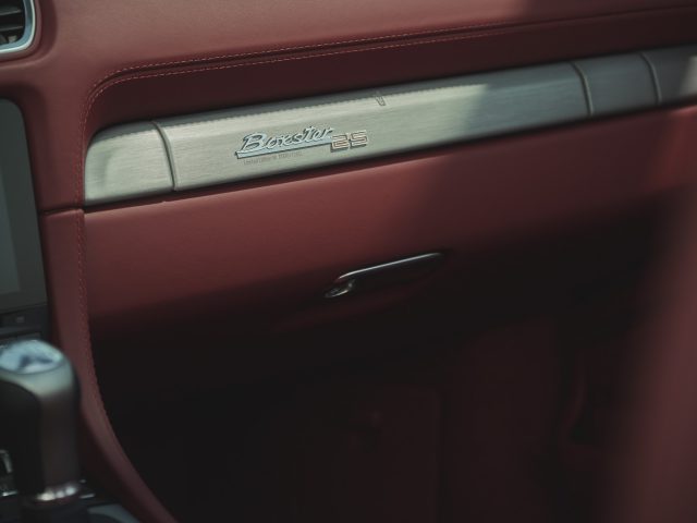 Close-up van het dashboard en het dashboardkastje in een auto met rood interieur, waarop trots een 'Boxster 25'-badge staat afgebeeld, wat de elegantie van de Porsche Boxster benadrukt.