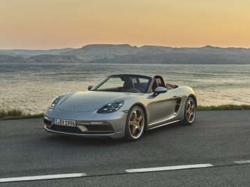 Een zilveren Porsche Boxster converteerbare sportwagen geparkeerd naast een meer tijdens zonsondergang, met heuvels op de achtergrond.