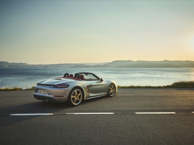 Een zilveren Porsche Boxster-cabriolet geparkeerd op een kustweg met de oceaan en de heuvels op de achtergrond tijdens zonsondergang.