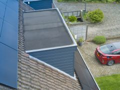 Een rode auto geparkeerd op een geplaveide oprit naast een huis met zonnepanelen op het dak, een voorbeeld van moderne Energievergelijken-opties. Groen omringt het gebied, wat bijdraagt aan de milieuvriendelijke charme.