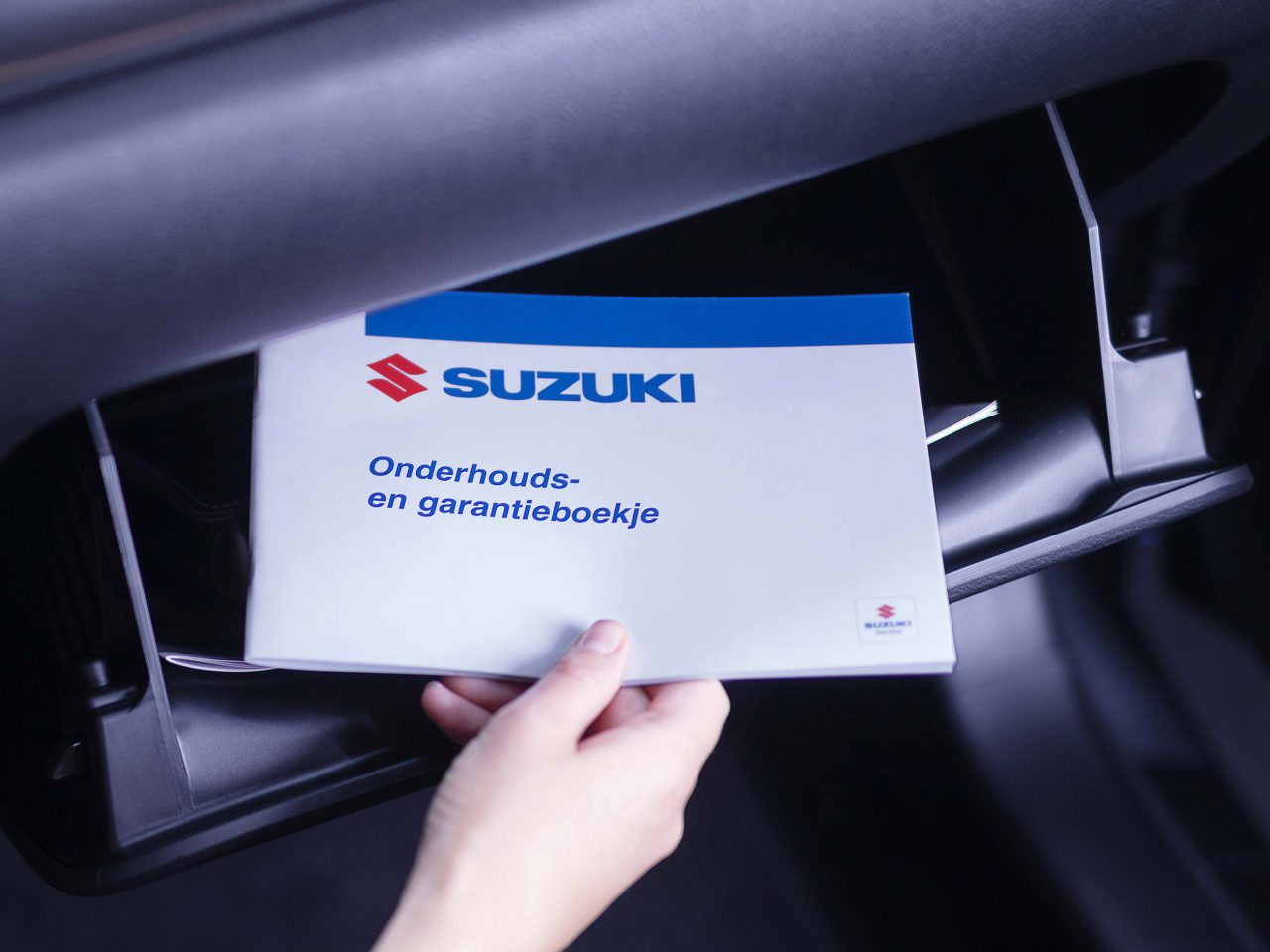 Een persoon die een Suzuki-onderhouds- en garantieboekje haalt uit het dashboardkastje van een voertuig.