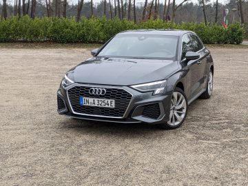 Een grijze Audi A3 TFSI e geparkeerd op een ruige ondergrond met op de achtergrond bomen en groen. Op het kenteken staat "IN-A 3254E.