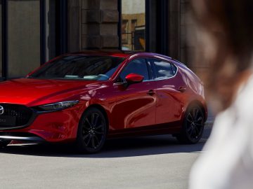 Een rode Mazda 3 hatchback staat op straat geparkeerd bij een gebouw. De onscherpe voorgrond onthult een gedeeltelijk zichtbare persoon.