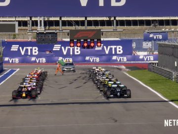 Een startgrid van raceauto's opgesteld op een circuit onder een groot spandoek met logo's, onder toezicht van marshals en met Netflix-branding zichtbaar in de rechteronderhoek, ter promotie van Formula 1 Drive to Survive seizoen 3.