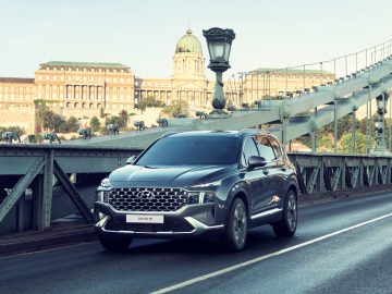 Een zilverkleurige Hyundai Santa Fe rijdt over een historische stalen hangbrug met een sierlijke lantaarnpaal, tegen de achtergrond van een ouder gebouw in Europese stijl.