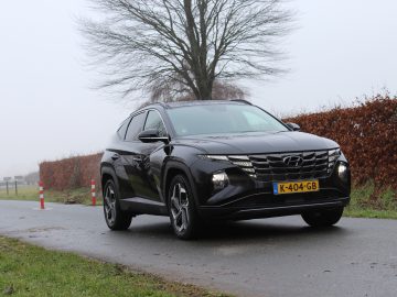 Een zwarte Hyundai Tucson SUV met kenteken K-404-GB staat geparkeerd op een smalle, mistige weg met een boom en een heg op de achtergrond. Aan de linkerkant van de weg zijn oranje en witte wegmarkeringen zichtbaar.