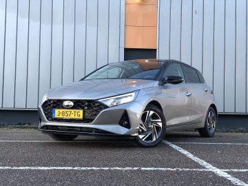 Een grijze Hyundai i20 hatchback staat geparkeerd op een asfaltoppervlak voor een modern gebouw met verticale metalen panelen. De auto heeft een geel kenteken met het nummer J-857-TG.