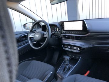 Binnenaanzicht van een moderne Hyundai i20 met het stuur, het dashboard met digitaal display, de versnellingspook en verschillende bedieningselementen. Het algehele kleurenschema is zwart met enkele metallic accenten.