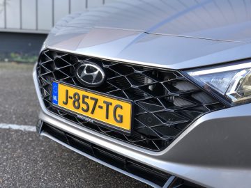 Close-up van de voorkant van een grijze Hyundai i20 met een Europees kenteken met de tekst "J-857-TG" uit Nederland. Op de achtergrond is een deel van een gebouw te zien.