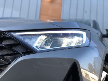Close-up van de koplamp van een Hyundai i20, verlicht met LED-verlichting, waarbij een deel van de slanke grille en de metallic grijze carrosserie zichtbaar zijn.