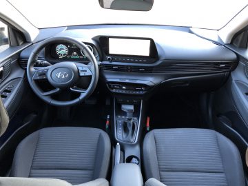 Het interieur van de Hyundai i20 getuigt van een strak, modern ontwerp met een stuurwiel, digitaal dashboard, centraal touchscreen, versnellingspook en twee voorstoelen versierd met grijze bekleding.