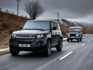 Twee Land Rover Defenders, een moderne en een klassieke, rijden op een landelijke weg met een heuvelachtig, bewolkt landschap. Het gebrul van de Land Rover Defender V8 galmt door het serene landschap en combineert erfgoed en kracht naadloos.