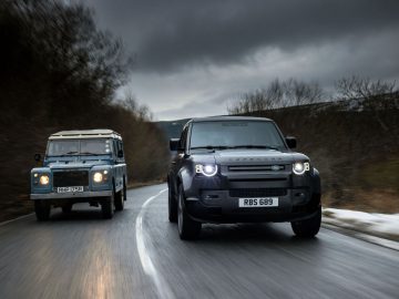 Twee Land Rover-voertuigen rijden over een natte, bochtige weg met aan weerszijden kale bomen. Het voertuig aan de linkerkant is een ouder, blauw model, terwijl het voertuig aan de rechterkant een strakke Land Rover Defender V8 is in het zwart. De lucht is bewolkt.