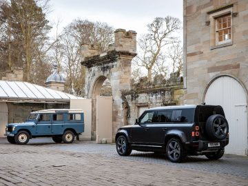 Twee Land Rovers geparkeerd op een stenen binnenplaats: een ouder blauw model en een moderne zwarte Land Rover Defender V8, tegen een achtergrond van historische stenen gebouwen met grote bogen en bomen op de achtergrond.