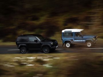 Twee SUV's, een zwarte en een blauwe met een wit dak, rijden zij aan zij op een weg door een bosrijke omgeving. De Land Rover Defender V8-motoren zijn in beweging, met een enigszins onscherpe achtergrond.