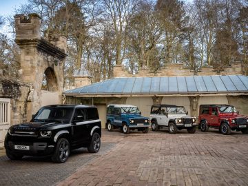 Vier Land Rover Defender V8-voertuigen staan geparkeerd voor een historisch gebouw met bomen op de achtergrond. De voertuigen zijn zwart, blauw, zilver en rood.