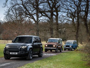 Drie Land Rover-voertuigen, waaronder een moderne zwarte Land Rover Defender V8 en twee klassieke modellen in bruin en blauw, rijden op een smal pad omringd door bomen en grasvelden.