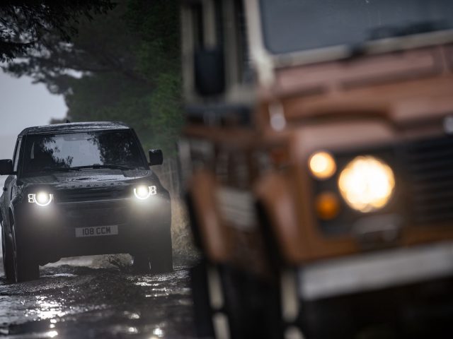 Twee terreinwagens in een bosrijke omgeving, één scherp in beeld met koplampen aan op de achtergrond en de andere gedeeltelijk zichtbaar en onscherp op de voorgrond. De grond is modderig en nat, perfect terrein voor een Land Rover Defender V8 om zijn robuuste capaciteiten te demonstreren.