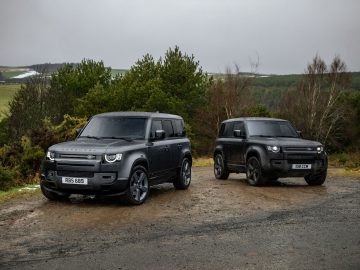 Twee zwarte SUV's, waaronder een strakke Land Rover Defender V8, staan geparkeerd op een natte, modderige weg met bomen en een verre, bewolkte landschap op de achtergrond.