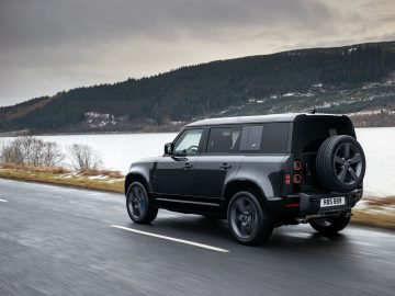 Een Land Rover Defender V8 rijdt op een natte weg langs een meer met beboste heuvels op de achtergrond onder een bewolkte hemel.