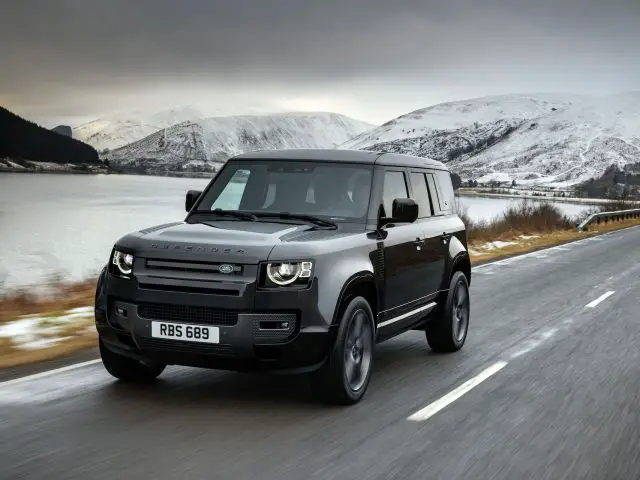 Een zwarte Land Rover Defender V8 rijdt over een weg langs een besneeuwd landschap met bergen en een meer op de achtergrond.