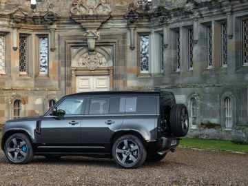 Een strakke Land Rover Defender V8 staat geparkeerd voor een oud stenen gebouw met ingewikkelde architectonische details en meerdere ramen.