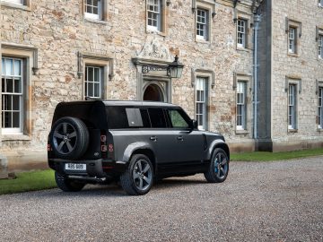 Een Land Rover Defender V8 staat geparkeerd op een grindpad naast een historisch stenen gebouw met grote ramen en een sierlijke ingang.