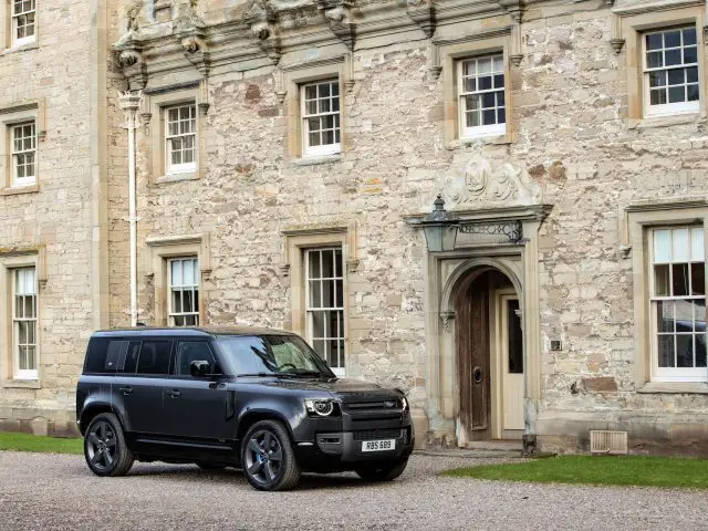 Een Land Rover Defender V8 staat geparkeerd voor een historisch stenen gebouw met meerdere ramen en een decoratieve ingang.