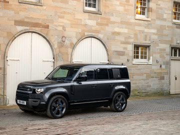 Een zwarte Land Rover Defender V8 staat geparkeerd in een geplaveide straat voor een stenen gebouw met gebogen houten deuren en kleine ramen.